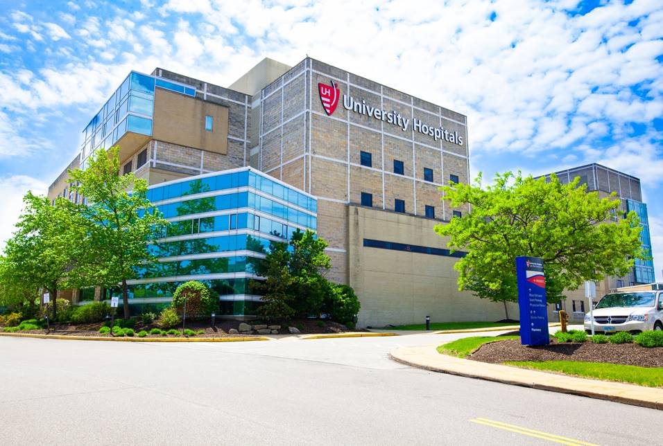 Multi-story University Hospitals building with brick and glass facade, blue wayfinding sign at entrance drive.