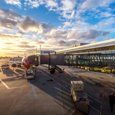 Commercial aircraft docked at terminal gate on airport tarmac at sunset, with ground crew vehicles and passengers nearby.