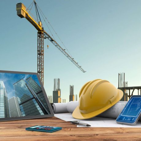 Yellow hard hat, rolled blueprints, tablet, and smartphone on a wooden desk with a construction site and tower crane in the background.
