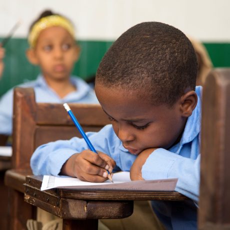 Young student in blue uniform writing in a notebook at a wooden school desk, with classmates in the background.