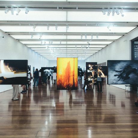 Large gallery hall with polished wood floors, white walls, large photographic prints on stands, and visitors browsing the exhibition.