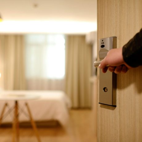 Hand gripping a lever door handle to open a hotel room door, with a bed and warm lighting visible inside.