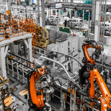 Automotive assembly line with multiple orange robotic arms welding car body frames inside a large industrial facility.