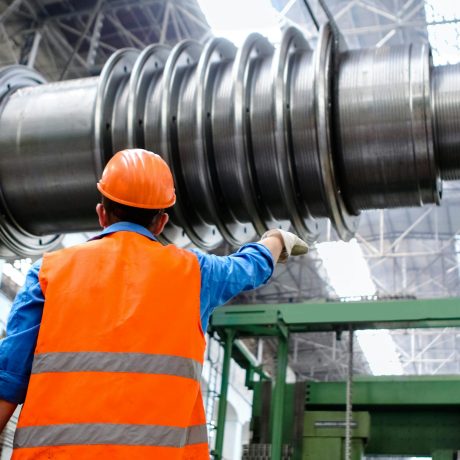 Worker in orange hi-vis vest and hard hat inspecting a large industrial rotor shaft inside a manufacturing facility.