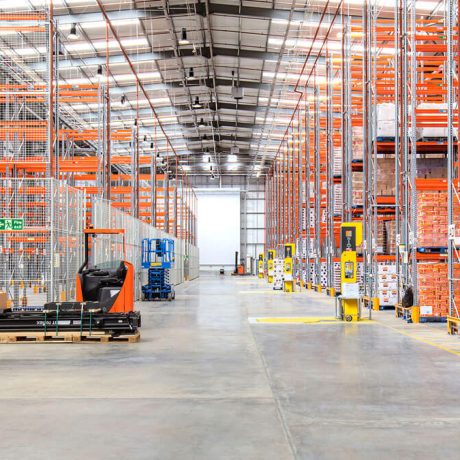Wide warehouse aisle with tall orange pallet racking, loaded shelves, a ride-on pallet truck, and a scissor lift on a concrete floor.